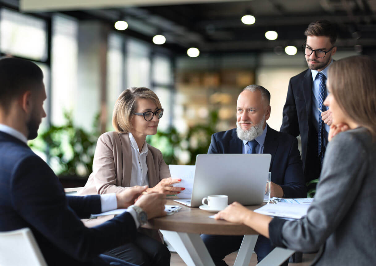Grupo de executivos em reunião corporativa, reunidos ao redor de uma mesa com notebook e documentos, discutindo estratégias em ambiente profissional moderno.
