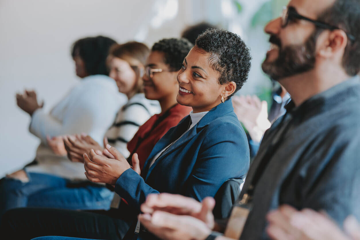 Pessoas sentadas em uma palestra aplaudindo, com expressões de atenção e satisfação, representando um ambiente corporativo ou evento de capacitação.