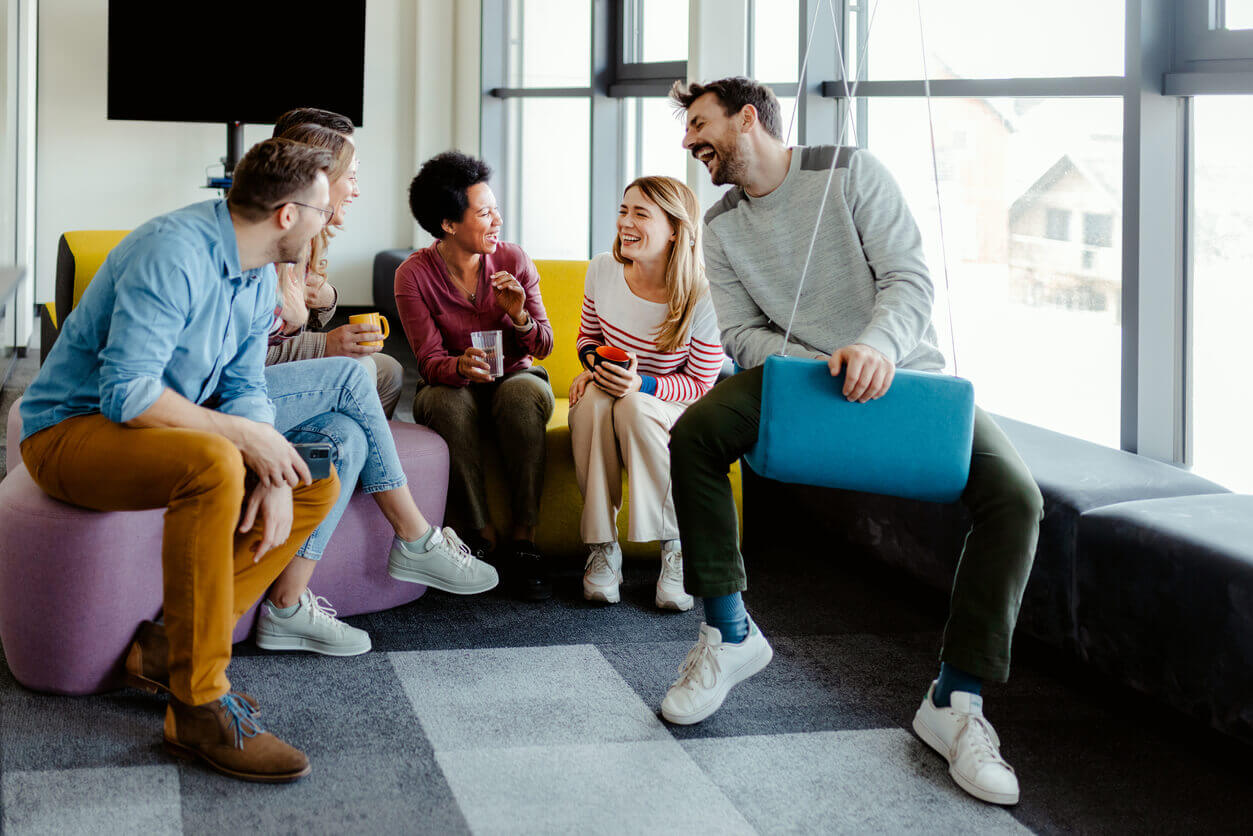Grupo de colegas de trabalho sorrindo e conversando descontraidamente em um ambiente corporativo moderno, representando bem-estar e ações de saúde mental no ambiente profissional.