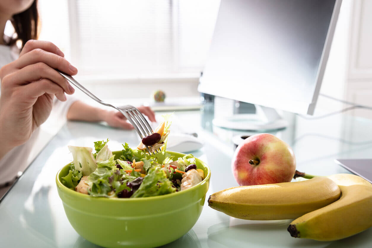 Pessoa comendo salada fresca ao lado de frutas no ambiente de trabalho, ilustrando hábitos saudáveis promovidos por consultoria nutricional para empresas.