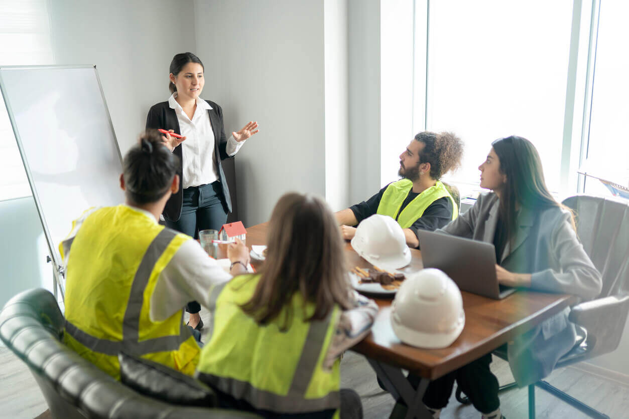 Reunião de equipe com profissionais de segurança do trabalho usando coletes refletivos, capacetes sobre a mesa e uma mulher apresentando informações em um quadro branco, ilustrando a aplicação do PGR conforme NR-1 na prática empresarial.