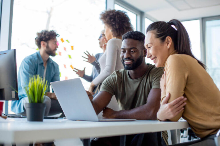 Colegas de trabalho sorrindo diante de um notebook em ambiente colaborativo, representando ações da NR1 para prevenção de riscos psicossociais nas empresas.
