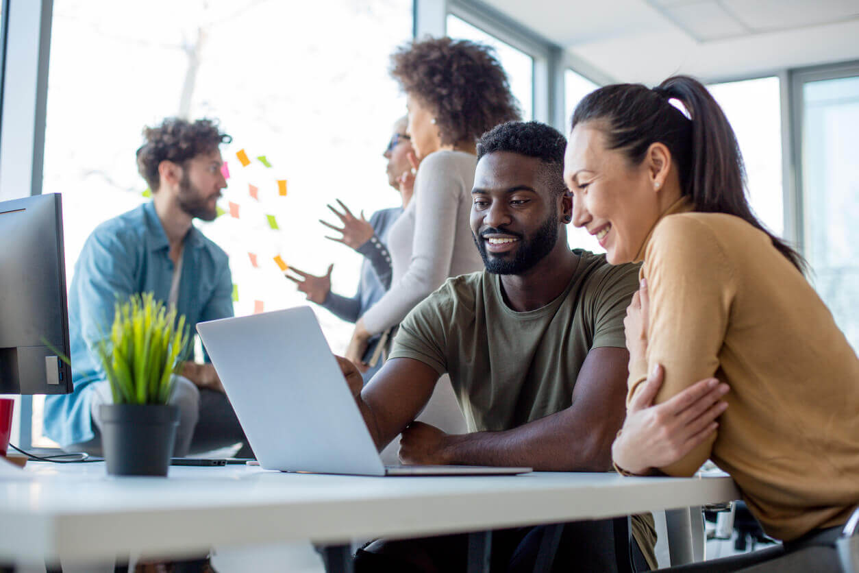 Colegas de trabalho sorrindo diante de um notebook em ambiente colaborativo, representando ações da NR1 para prevenção de riscos psicossociais nas empresas.